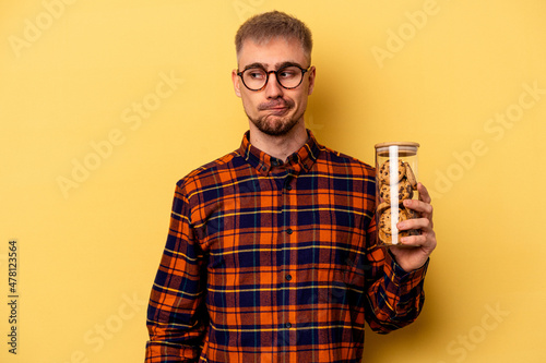 Young caucasian man holding cookies jar isolated on yellow background confused, feels doubtful and unsure Fotobehang
