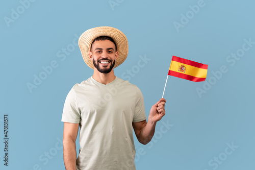 Happy arab man wearing t-shirt and hat, holding the spanish flag and smiling at camera, standing over blue background