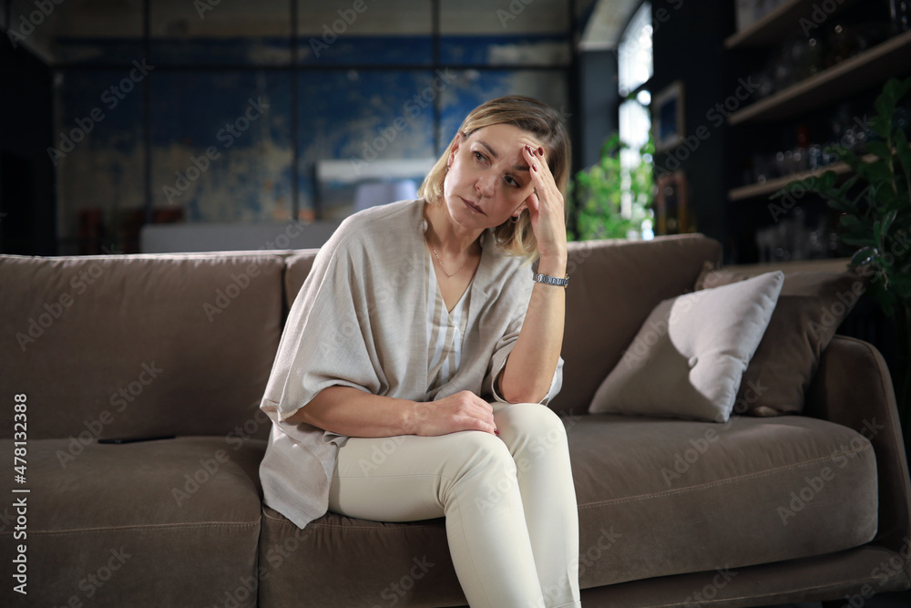 Fototapeta premium Stressed middle aged woman sit on sofa in living room, lost in thought.