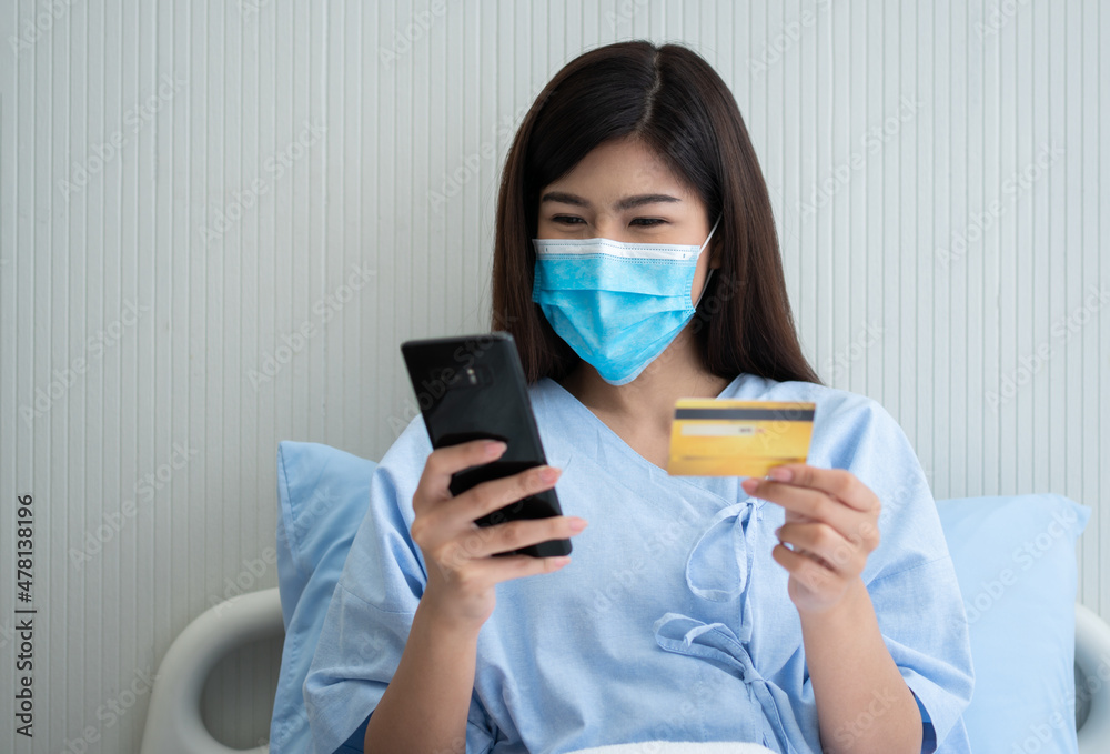 Happy Asian woman wearing a medical mask and holding mock up credit /insurance card and smartphone in a hospital bed for Check health insurance eligibility. Insurance policy by bank, payment medical Happy Asian woman wearing a medical mask and holding mock up credit /insurance card and smartphone in a hospital bed for Check health insurance eligibility. Insurance policy by bank, payment medical