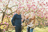 Outdoor portrait of happy young family playing in spring park under blooming magnolia tree, lovely couple with two little children having fun in sunny garden