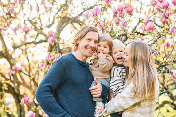 Outdoor portrait of happy young family playing in spring park under blooming magnolia tree, lovely couple with two little children having fun in sunny garden