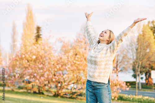 Outdoor portrait of young happy blond woman in spring park, emotional female model having fun on sunny warm day, pulling arms up wide open