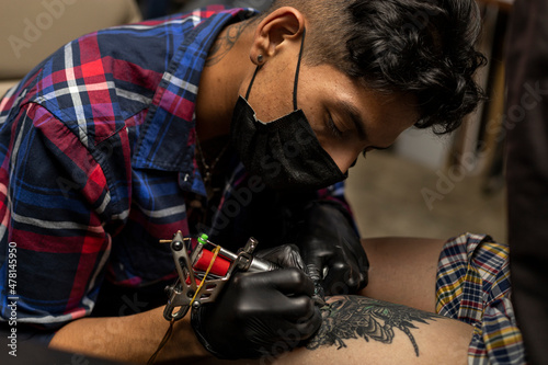 Venezuelan tattoo artist with mouth cover and black gloves performing a tattoo created by him. Body art concept