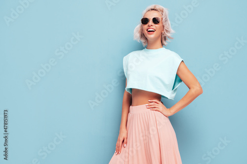 Young beautiful smiling female in trendy summer clothes. Sexy carefree woman posing near blue wall in studio. Positive blond model having fun and going crazy. In sunglasses