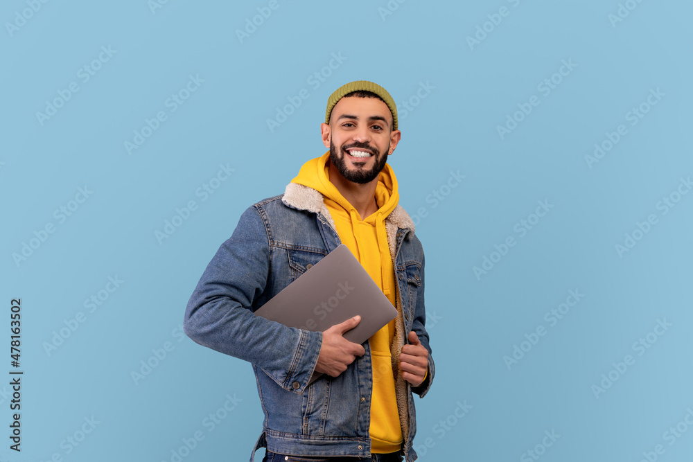 Naklejka premium Happy middle eastern guy holding laptop and smiling at camera, arabic man with pc computer over blue background