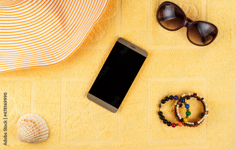 Women's accessories for a beach holiday. A smartphone with a blank screen, a striped hat, jewelry, sunglasses, and a shell in the background of a yellow terry towel.