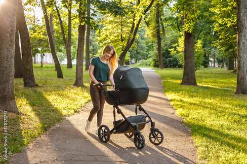 Young blonde woman walking with black stroller in summer park. Happy mother with baby in pram outdoors.  photo