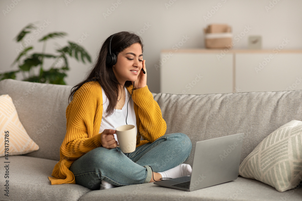 Telecommuting concept. Arab woman sitting on sofa and working online on laptop, wearing headset and holding cup Telecommuting concept. Arab woman sitting on sofa and working online on laptop, wearing headset and holding cup