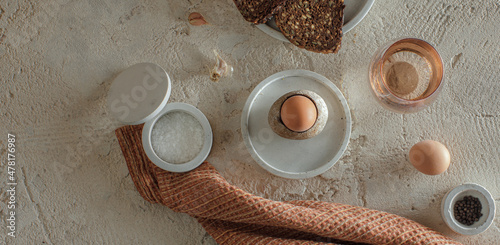 Breakfast setting with boiled egg in stoneware egg cup, whole grain rye bread, glass of water, salt flakes and pepper in concrete bowls on rough textured clay background. Healthy breakfast concept photo