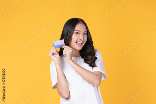 Portrait of a happy young asian woman wearing white shirt holding bank card, credit card isolated on yellow background. Business online shopping concept.
