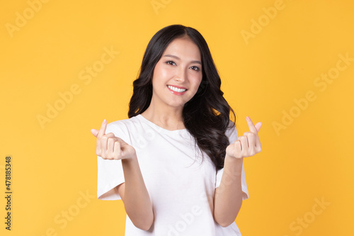 Portrait of beautiful young Asian woman model long hair in casual white shirt smile, happy and enjoying in studio isolated on yellow background. The model raises her hand as an heart symbol.