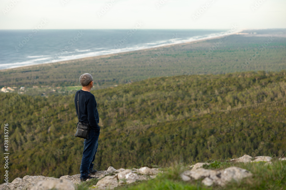 Thoughtful man with hands in his pockets,  standing on top of mountain,  staring at beautiful nature landscape. Forest and ocean background. copyspace. Inspiring and motivational view.