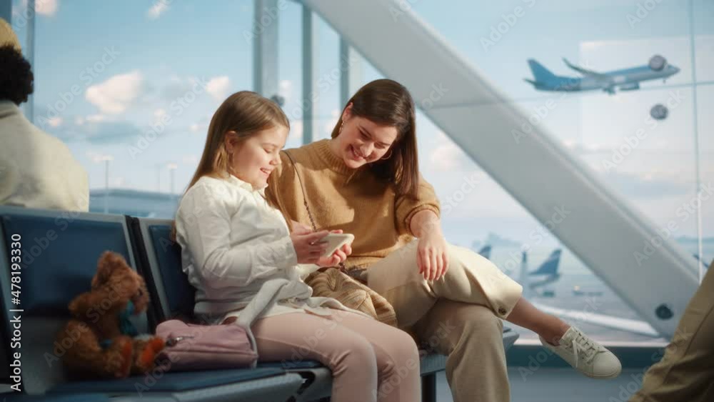 Busy Airport Airplane Terminal: Happy Beautiful Mother and Cute Little Daughter Wait for their Vacation Flight, Use Mobile Smartphone for Fun and Video Games. People in Boarding Lounge of Airline Hub