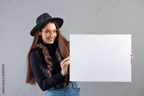elegant young woman in glasses and hat showing empty board banner, isolated on gray, copy space