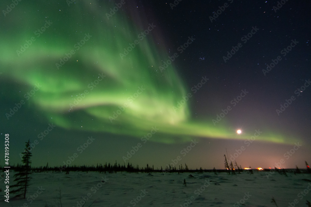 The northern lights and a rising moon fill the sky above a distant community in Canada's sub-arctic