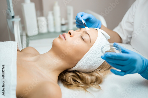 Hands of female cosmetologist applying a transparent mask with a brush on a female face . Facial skin peeling