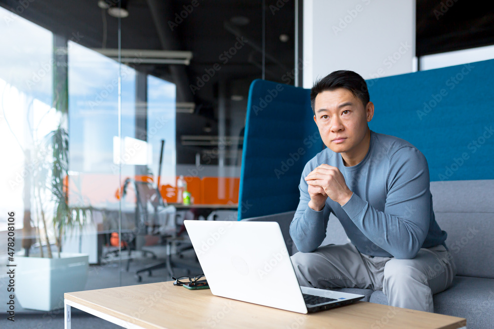A thinking Asian man works at a computer in a modern office, a freelancer is focused on solving a task, makes an important job. A thinking Asian man works at a computer in a modern office, a freelancer is focused on solving a task, makes an important job.