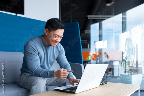 Happy Asian man shopping online, working in a modern office on a laptop, smiling and rejoicing, the chosen product, holding a credit card
