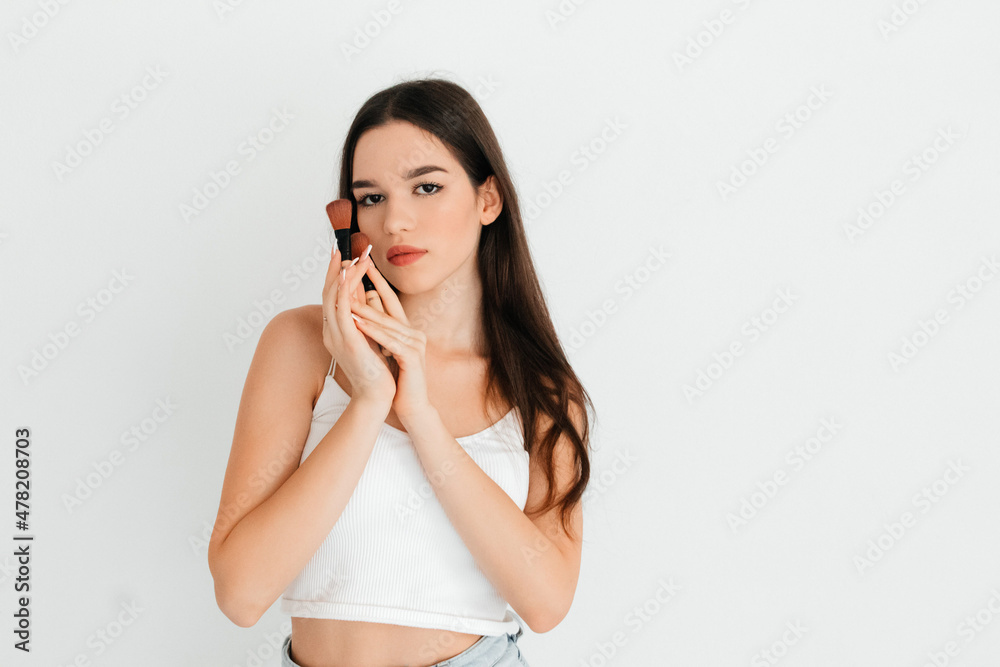 Young woman in the process of making makeup holding cosmetic brush near her face, posing on white studio background. Morning routine concept, getting ready for work Young woman in the process of making makeup holding cosmetic brush near her face, posing on white studio background. Morning routine concept, getting ready for work