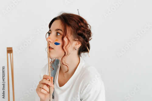 Portrait of thoughtful redhead painter with brush on white background. A professional young woman with paint on her cheek, searching for an inspiration in her work