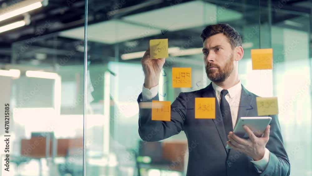 serious businessman analyst develops a strategy for enterprise, financial growth thinking make startup plan in office Business male employee in suit write on paper sticker sticky notes on glass wall