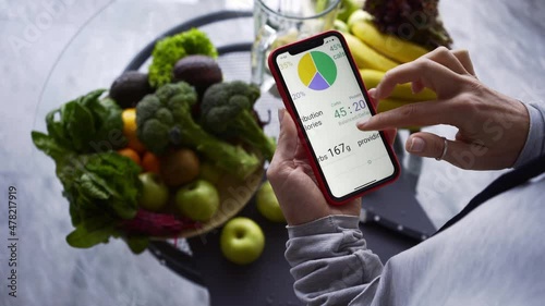 Unrecognizable lady uses application on smartphone to check calories of recipe on table with fresh fruits and vegetables in contemporary kitchen close view photo
