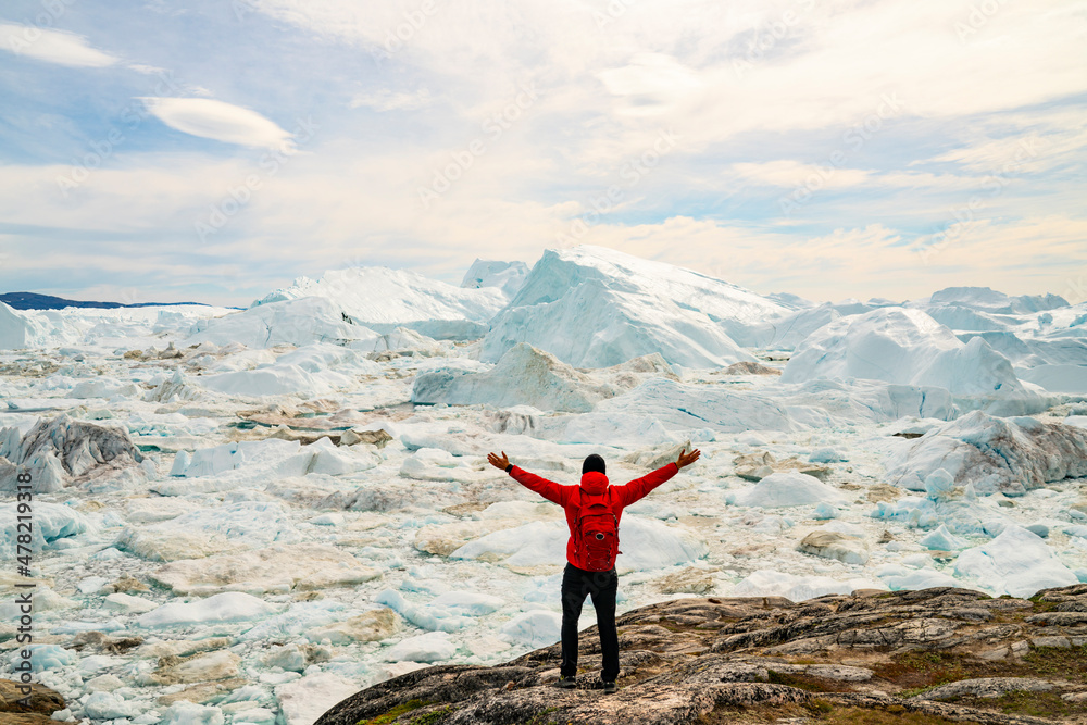 Obraz premium Climate Change and Global Warming Optimism and Positivity Concept. Man on Greenland arms raised in awe of iceberg nature. Melting of glaciers and Greenland ice sheet is a cause of sea levels rise