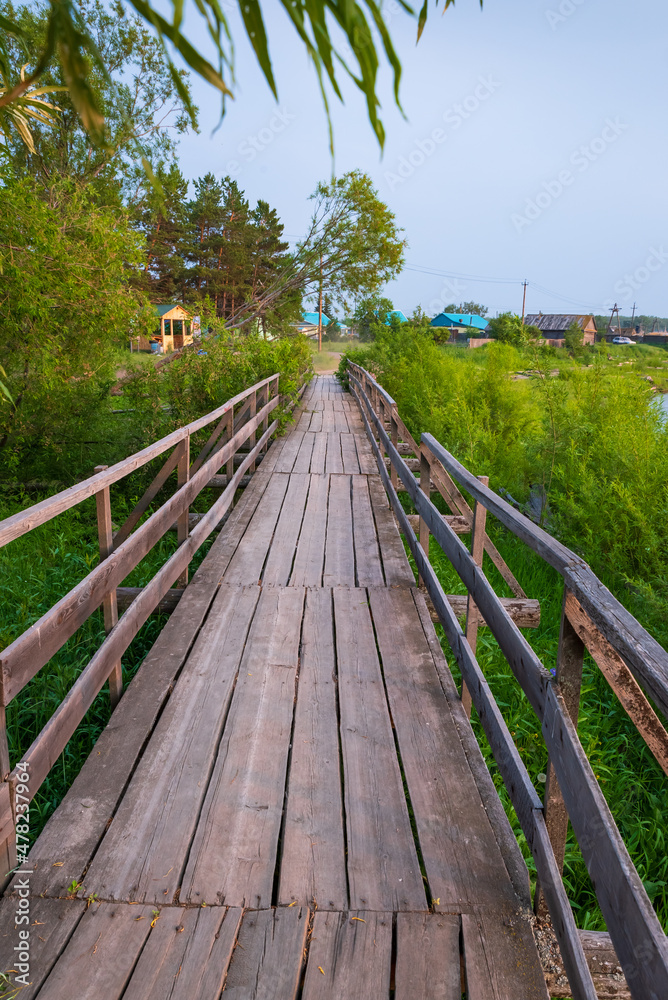 Fototapeta premium Wooden bridge in the village across the river, near grass, bushes and trees.