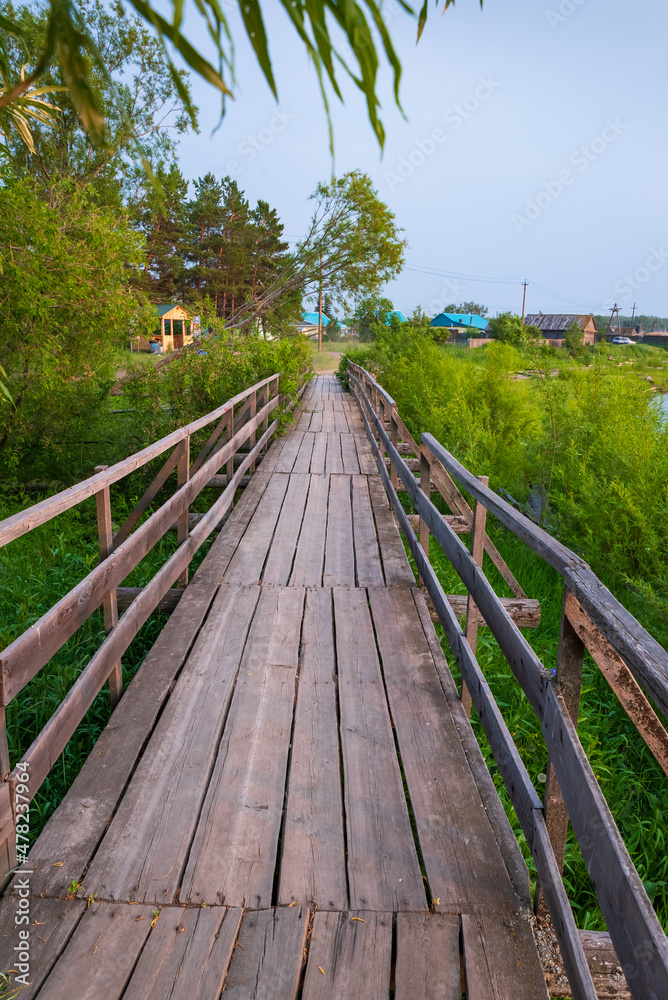 Wooden bridge in the village across the river, near grass, bushes and trees.