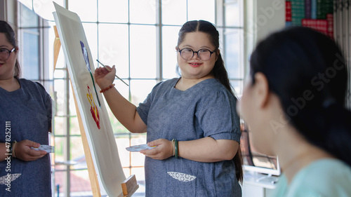 Foto Portrait shot of Asian happy lovely mother standing smiling with young chubby down syndrome autistic autism little daughter wearing eyeglasses holding paintbrush drawing painting on canvas easel