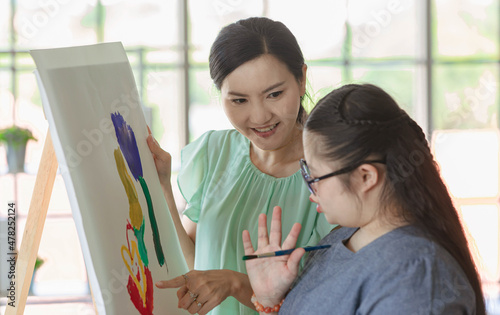 Close up shot of Asian happy lovely mother face standing smiling encouraging young chubby down syndrome autistic autism little daughter while using paintbrush drawing painting colors on canvas easel