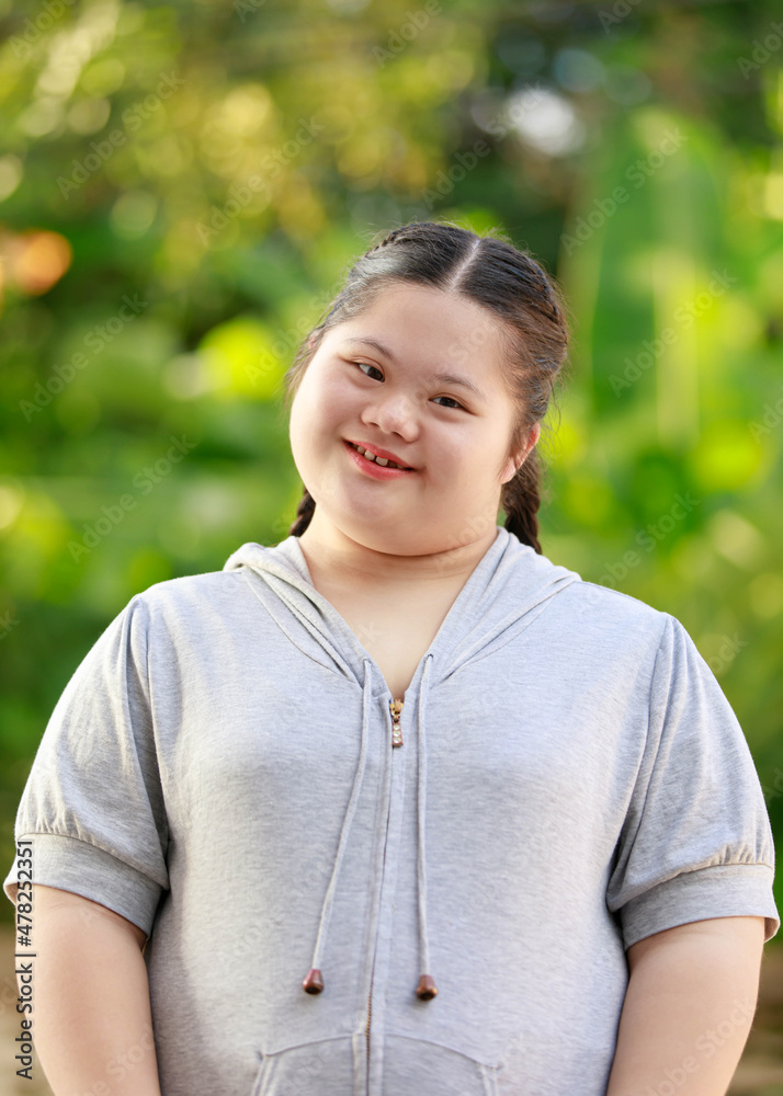 Portrait shot of Asian young chubby down syndrome autistic autism little cute schoolgirl with braid pigtail hairstyle model stand posing smiling look at camera in front blurred garden view background Portrait shot of Asian young chubby down syndrome autistic autism little cute schoolgirl with braid pigtail hairstyle model stand posing smiling look at camera in front blurred garden view background