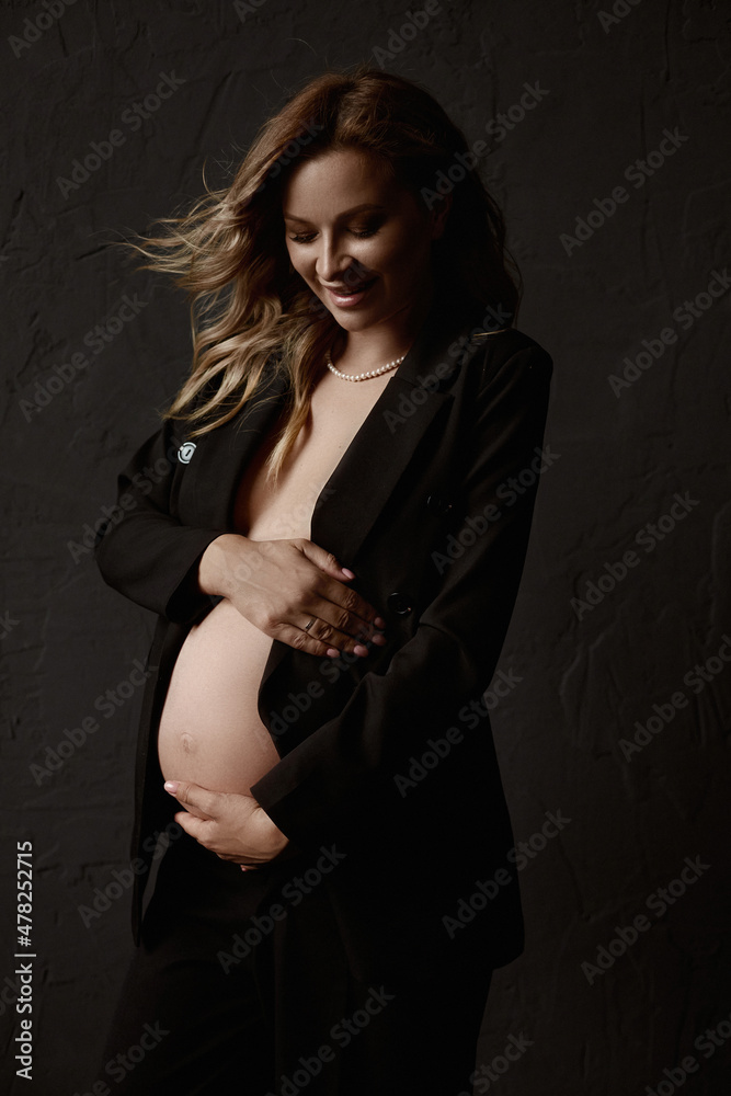 A gorgeous pregnant young woman with fluttering blond hair wearing an unbuttoned blazer embracing her belly and posing beside the black wall. Naked belly. Lifestyle, pregnancy, health A gorgeous pregnant young woman with fluttering blond hair wearing an unbuttoned blazer embracing her belly and posing beside the black wall. Naked belly. Lifestyle, pregnancy, health