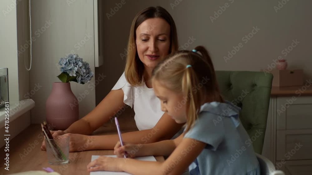 Young mother teacher gently tapping the little primary school girl child on back praising doing homework together, successfully doing test. Happy female tutor helping to daughter with homework. Young mother teacher gently tapping the little primary school girl child on back praising doing homework together, successfully doing test. Happy female tutor helping to daughter with homework.