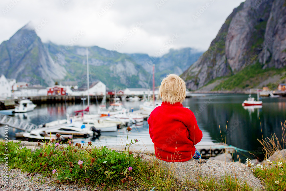 Cute child in red jacket, enjoying the view of a typical wooden red houses, called rorbuer in Lofoten, Norway