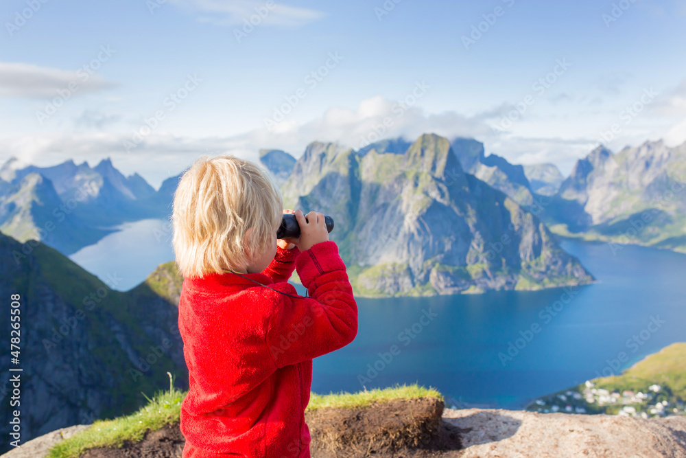 Cute child, standing on top of the mountains and looking down on Reine after climbing Reinebringen treeking path with lots of stairs, using binoculars