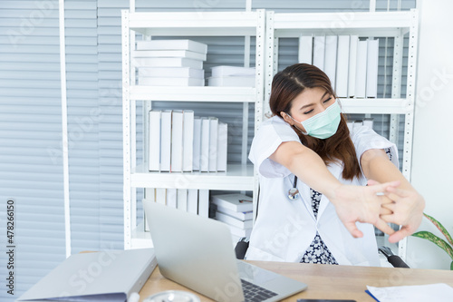 Portrait of young Asian female doctor with white coat is stretching her body while using computer laptop to check, diagnosis and parepare patient's medical information to treatment on desk in hospital photo