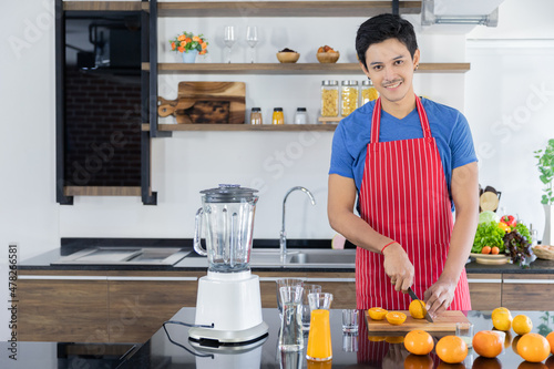Healthy handsome man with casual clothes is smiling and preparing orange juice to diet, in kitchen at home in holiday.