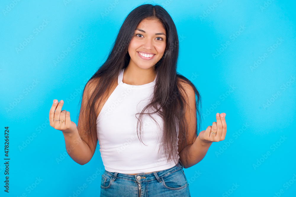 Young hispanic girl wearing tank top over blue background , making money gesture. Young hispanic girl wearing tank top over blue background , making money gesture.