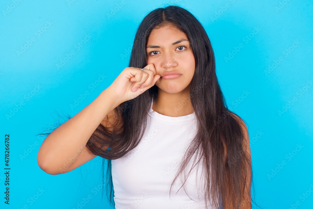 Unhappy Young hispanic girl wearing tank top over blue background crying while posing at camera whipping tears with hand. Unhappy Young hispanic girl wearing tank top over blue background crying while posing at camera whipping tears with hand.