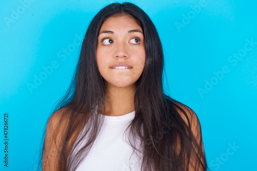 Young hispanic girl wearing tank top over blue background with thoughtful expression, looks away keeps hands down bitting his lip thinks about something pleasant.