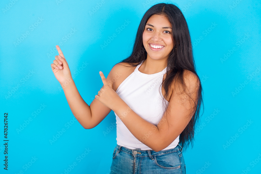 Positive Young hispanic girl wearing tank top over blue background with beaming smile pointing with two fingers and looking on empty copy space. Advertisement concept. Positive Young hispanic girl wearing tank top over blue background with beaming smile pointing with two fingers and looking on empty copy space. Advertisement concept.