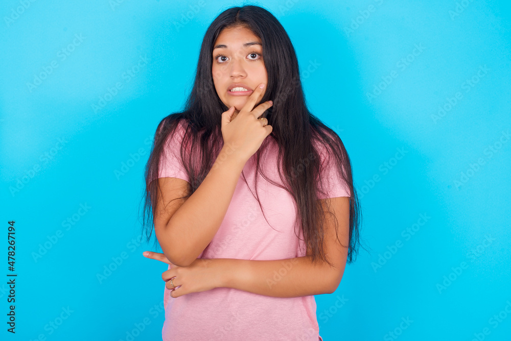 Young hispanic girl wearing pink T-shirt over blue background covering mouth with hands scared from something or someone bitting nails Young hispanic girl wearing pink T-shirt over blue background covering mouth with hands scared from something or someone bitting nails