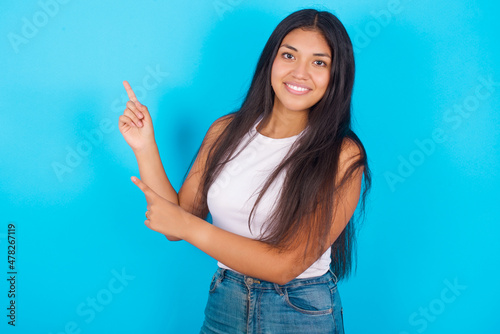 Young hispanic girl wearing tank top over blue background with positive expression, indicates with fore finger at blank copy space for your promotional text or advertisement.