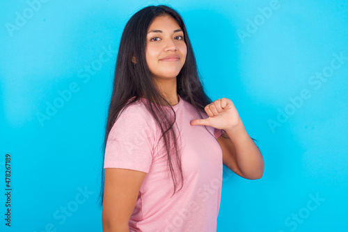 Closeup of cheerful Young hispanic girl wearing pink T-shirt over blue background looks joyful, satisfied and confident, points at himself with thumb.