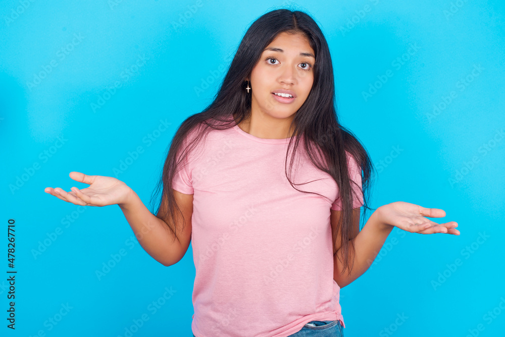 So what? Portrait of arrogant Young hispanic girl wearing pink T-shirt over blue background shrugging hands sideways smiling gasping indifferent, telling something obvious. So what? Portrait of arrogant Young hispanic girl wearing pink T-shirt over blue background shrugging hands sideways smiling gasping indifferent, telling something obvious.