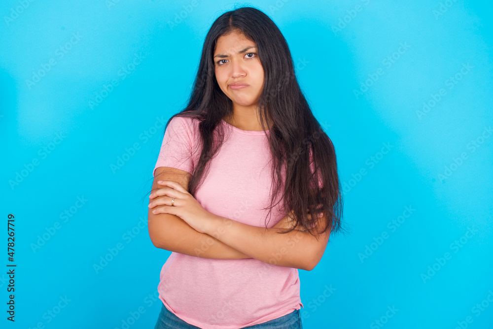 Picture of angry Young hispanic girl wearing pink T-shirt over blue background crossing arms. Looking at camera with disappointed expression. Picture of angry Young hispanic girl wearing pink T-shirt over blue background crossing arms. Looking at camera with disappointed expression.