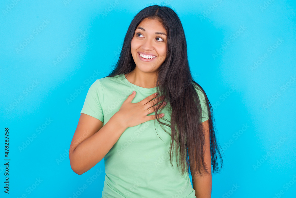 Joyful Young hispanic girl wearing green T-shirt over blue background expresses positive emotions recalls something funny keeps hand on chest and giggles happily. Joyful Young hispanic girl wearing green T-shirt over blue background expresses positive emotions recalls something funny keeps hand on chest and giggles happily.