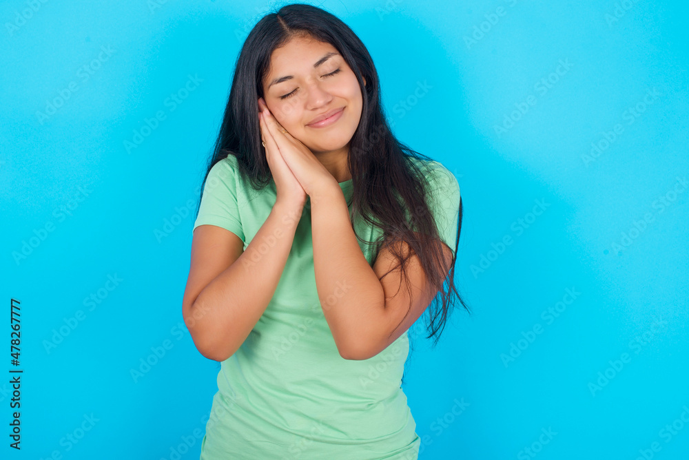 Young hispanic girl wearing green T-shirt over blue background leans on pressed palms closes eyes and has pleasant smile dreams about something Young hispanic girl wearing green T-shirt over blue background leans on pressed palms closes eyes and has pleasant smile dreams about something
